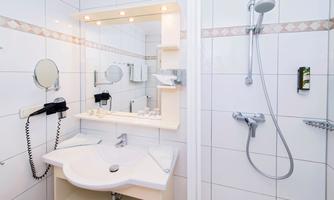 A modern bathroom with white tiles and a large mirror. There is a shower, a sink, and various bathroom utensils.
