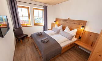 A cozy bedroom with two single beds and warm wooden details. Large windows let in plenty of daylight.