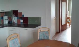 A modern kitchen with a wooden table and chairs. In the background, another room with natural light is visible.
