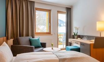 A cozy room with a double bed and a chair. The view leads to a balcony with a beautiful view of the greenery.