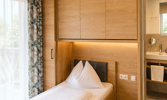 A cozy bedroom with a single bed and wooden shelves. The room has large windows that let in a lot of light.
