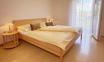 A cozy bedroom with a double bed and soft pillows. The room has large windows with curtains and a small table with a lamp.