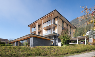 A modern, multi-story building with large windows and balconies. Surrounded by green grass and trees in a mountainous landscape.