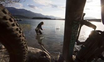 A person is standing in the water and examining something. In the foreground, bicycle tires are visible, while the landscape in the background features mountains and water.