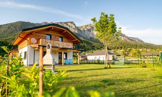 A beautiful house with wooden cladding in a green environment. In the background, mountains and a clear sky are visible.