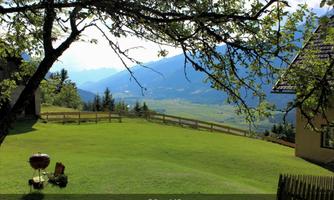 A green lawn with a grill and a chair, surrounded by mountains. The landscape is sunny and inviting.