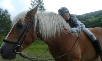 A girl hugs a horse and smiles. In the background, there are trees and a green meadow.