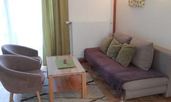A cozy living room with a gray sofa and two beige armchairs. The room is bright and cheerful, with a wooden table and a green curtain.