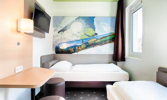 A modern hotel room with a single bed and an additional fold-out bed. A large painting hangs on the wall and a television is mounted.