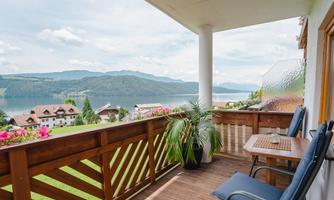 A cozy balcony with two chairs and a table. In the background, there is a beautiful view of the lake and the surrounding mountains.