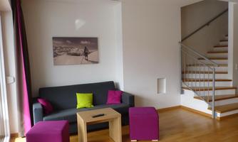 A modern living room with a gray sofa and colorful cushions. In the background, there is a staircase and a piece of artwork on the wall.