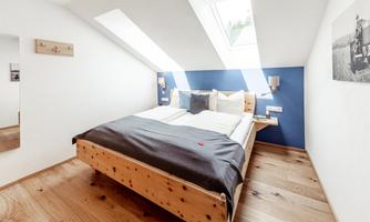 A modern bedroom with a wooden double bed and a blue accent wall. Large windows provide plenty of daylight and a bright atmosphere.