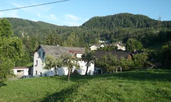 A picturesque house surrounded by green meadows and trees. In the background, gentle hills and a clear blue sky can be seen.