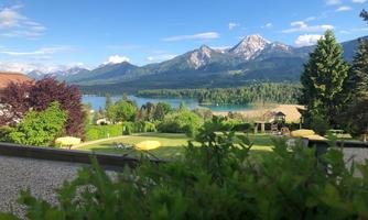 A beautiful view of a lake, surrounded by mountains and green forest. The sky is blue and clear, highlighting the natural beauty of the landscape.