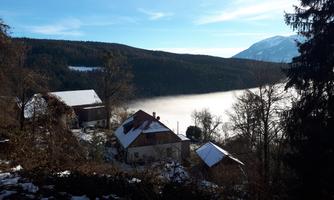 A picturesque landscape with snow-covered houses and a misty valley. In the background, forests and mountains are visible under a clear blue sky.