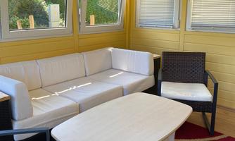 A cozy interior with white sofas and a small table. The walls are painted yellow and there are large windows that let in plenty of light.
