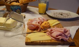A breakfast table with a wooden top displaying various types of sausages and cheeses. In the background, rolls, jam, and a glass of orange juice are visible.