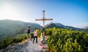 A family stands at a mountain summit with a wooden cross. In the background, green hills and a clear sky can be seen.