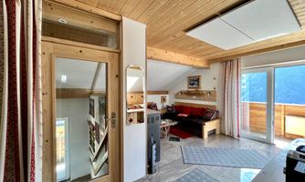 A cozy living space with wooden paneling and large windows. The room is bright and inviting, with a terrace offering a view of the mountains.