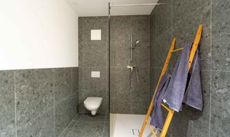 A modern bathroom with gray tiles and a glass shower partition. A towel hangs on a wooden dryer next to a toilet.