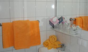 A modern bathroom with white tiles and orange towels. Various toiletries and glasses are visible on the sink.