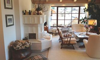 A cozy living room with a fireplace, comfortable armchairs, and large windows. There are appealing decorations and a warm, inviting style.
