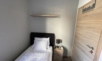 A simple room with a single bed, a nightstand, and a wooden shelf. The walls are painted in a neutral gray, and the door features a modern wood design.