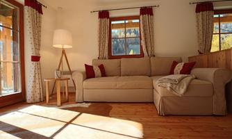 A cozy living room with a beige sofa and red cushions. Natural light streams through the windows, creating a warm atmosphere.