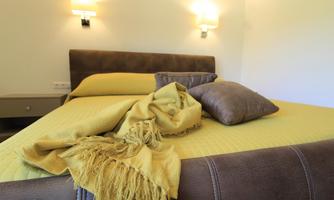 A cozy bedroom with a comfortable bed and a warm yellow blanket. The walls are light, and soft light radiates from the wall lights.