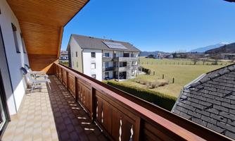 A spacious balcony with a wooden railing and a view of a green meadow. In the background, there are mountains and a blue sky.