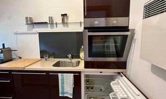 A modern kitchen with dark wooden cabinets and a compact oven. The sink and a refrigerator are also visible.