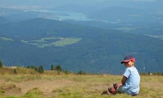 A child sits on a meadow and looks at a green hilly landscape. In the background, mountains and a lake are visible.