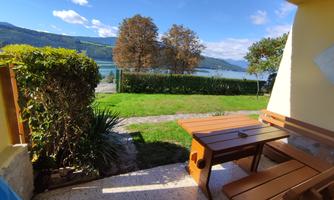 A cozy terrace with wooden furniture and a view of a lake. In the background, trees and mountains can be seen.
