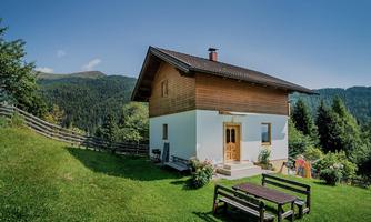 A cozy house on a meadow surrounded by trees and mountains. There is a table with benches in the foreground.