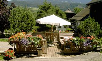 Cozy terrace with many colorful flowers and a sunshade. In the background, mountains and a green landscape are visible.