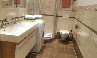 A modern bathroom with a shower, a sink, and a washing machine. The room has bright tiles and good lighting.