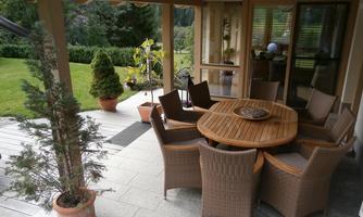 A cozy outdoor area with a round wooden table and chairs. In the background, a well-kept garden and a terrace are visible.