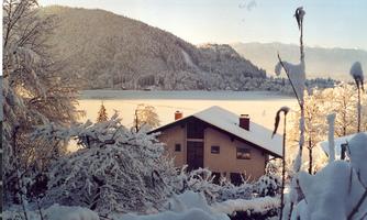 A snow-covered landscape with a house at the edge of a frozen lake. In the background, tall mountains and a clear sky can be seen.
