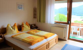 A cozy bedroom with a double bed and colorful pillows. Large windows provide a beautiful view of nature outside.