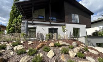 A modern house with a dark facade and large windows. Surrounded by a landscaped garden featuring stones and plants.