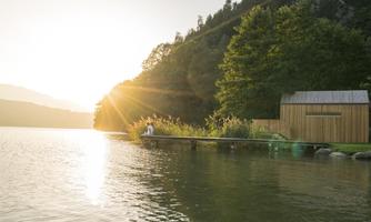 A calm lake at sunset with a wooden dock. In the background, there is a modern, small building surrounded by trees.