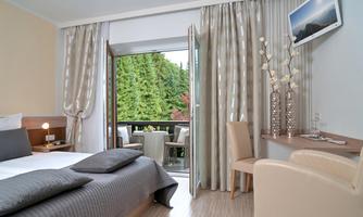 A modern hotel room with a bed and a desk. The windows open to a balcony with a view of a green environment.
