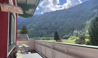 A picturesque mountain landscape with green hills and a blue sky. Small tables are on the veranda and flowers can be seen.
