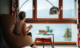 A person is sitting in a modern armchair, enjoying a view of the landscape through a large window. In front of them, books are on a table and there is a stylish lamp.
