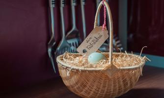 A basket with hay and an egg, decorated with a card. In the background, kitchen utensils are visible.