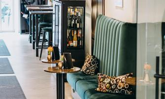 An elegant lounge with a green sofa and stylish cushions. In the background, tables and a lit drink wall can be seen.