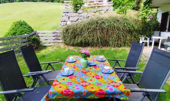 An inviting table with a colorful tablecloth is set in the garden. Surrounding the table are several chairs, and the landscape is green with a view of hills.