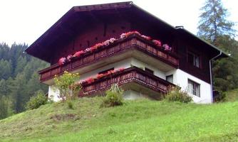 A traditional chalet in the mountains, surrounded by meadows and trees. The balcony is adorned with colorful flowers.
