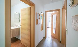 A bright corridor with wooden doors and tiled flooring. On the left side, there is a bathroom with a mirror.