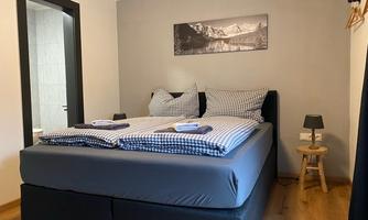 A modern bedroom with a double bed and checkered bedding. In the background, a landscape painting and a small nightstand can be seen.